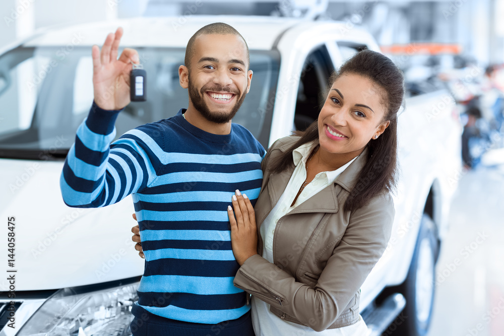 Smiling couple stands next to a white car, man holding up car key to celebrate a new vehicle.