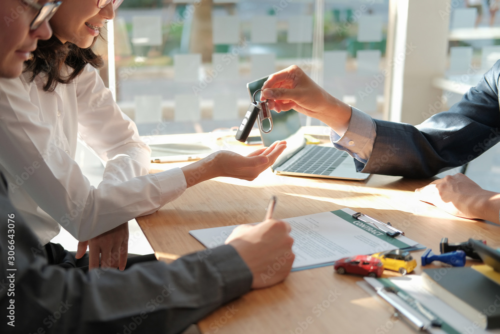Person hands over car keys to another person across a bright office table; a laptop and papers are on the desk.