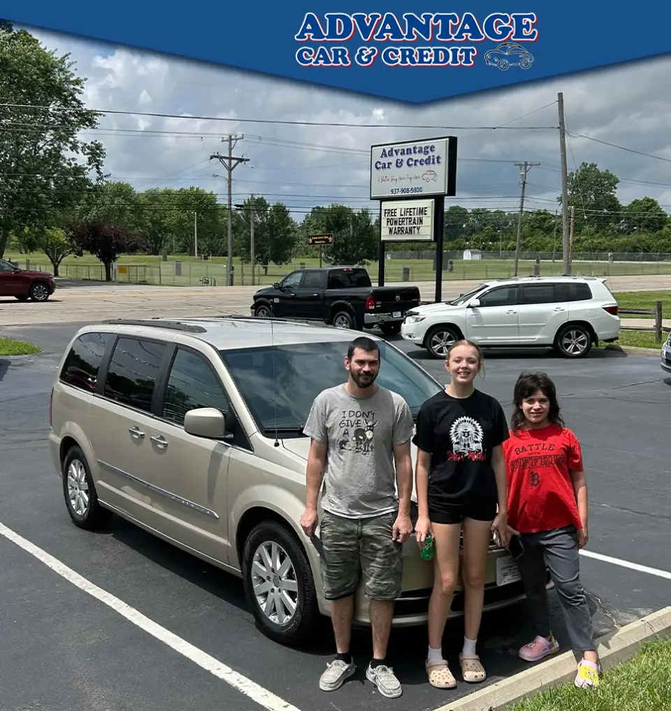 Three people stand beside a beige minivan in a parking lot, with an Advantage Car & Credit banner above them.