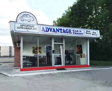 Front view of Advantage Car & Credit auto dealership storefront with glass doors, red curb, and trees in the background.
