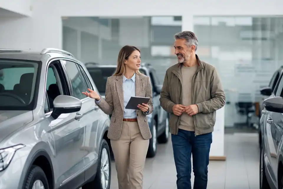 Saleswoman in a blazer explains features to a male customer beside a silver SUV in a bright car showroom.