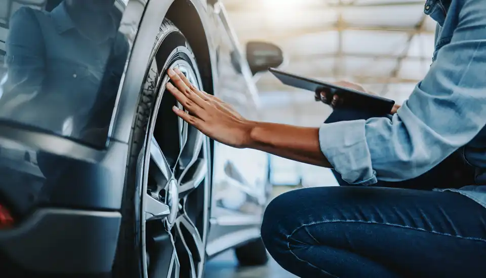 Person kneeling beside a car, inspecting the front left tire while holding a clipboard in a workshop setting
